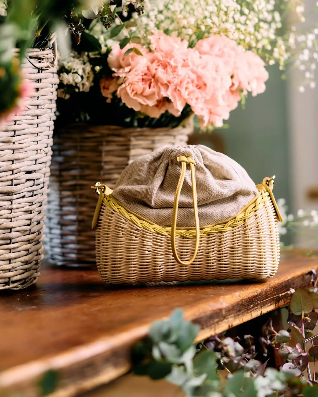 Straw pouch bag with a gold chain beside a basket of pink flowers