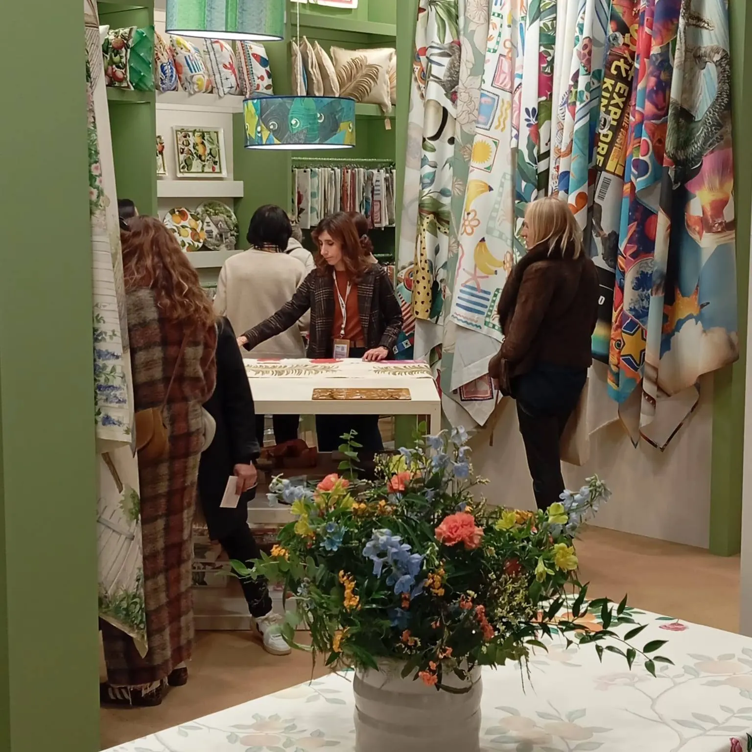 Visitatori allo stand fra i tessuti appesi, mazzo di fiori in primo piano
